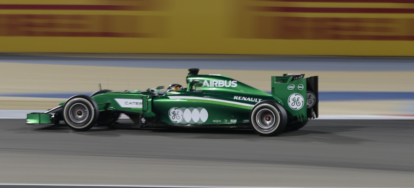 Green Caterham CT05 under floodlights at speed with a blurred yellow and red barrier behind