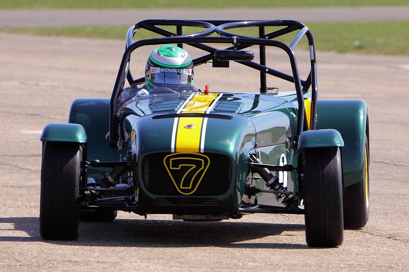 Green Caterham Seven with a yellow stripe and a 7 badge on its nose, driven by a man in a green Formula 1 helmet