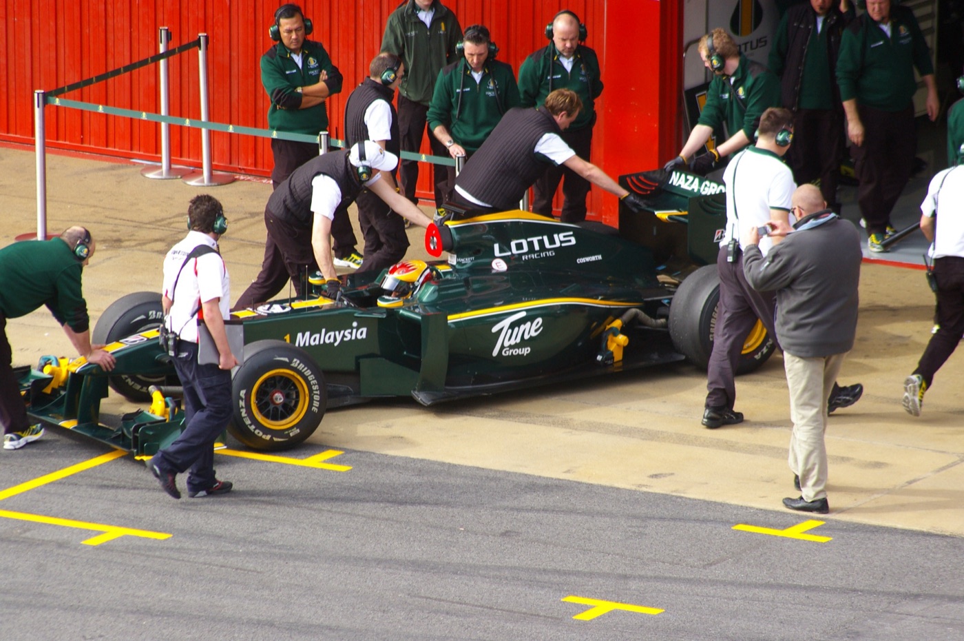 Green and yellow Lotus T127 being pushed into the pit garage by mechanics in green team jackets at a pre-season test