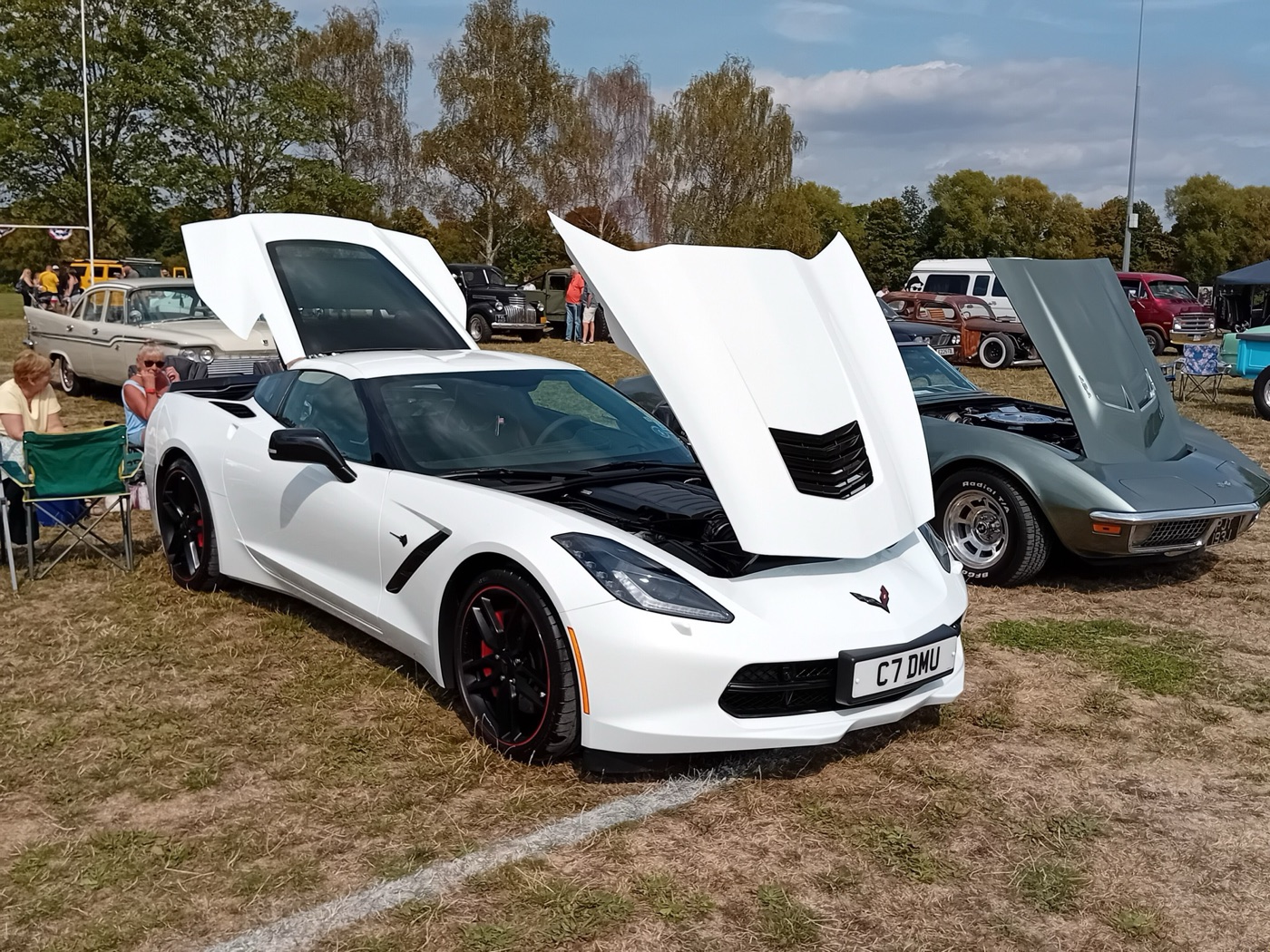 White C7 Corvette Stingray with British number plate, bonnet and roof panel open, at a grass-field car show next to a C3 Corvette
