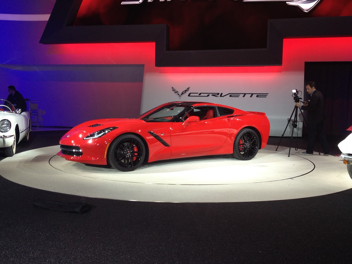 Red C7 Corvette Stingray coupe on a turntable under the Corvette crossed-flags sign, with a 1953 Corvette at the left edge