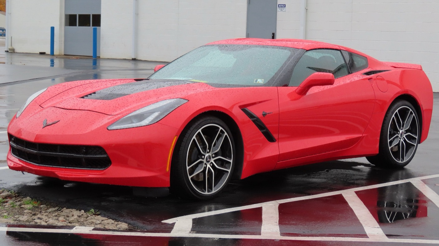 Red C7 Corvette Stingray coupe with a black bonnet vent and split-spoke wheels, parked on a wet forecourt