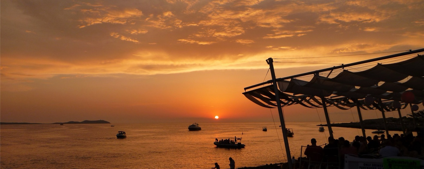 Sun setting into the sea off Ibiza with boats on the water and diners under the awnings of Café del Mar