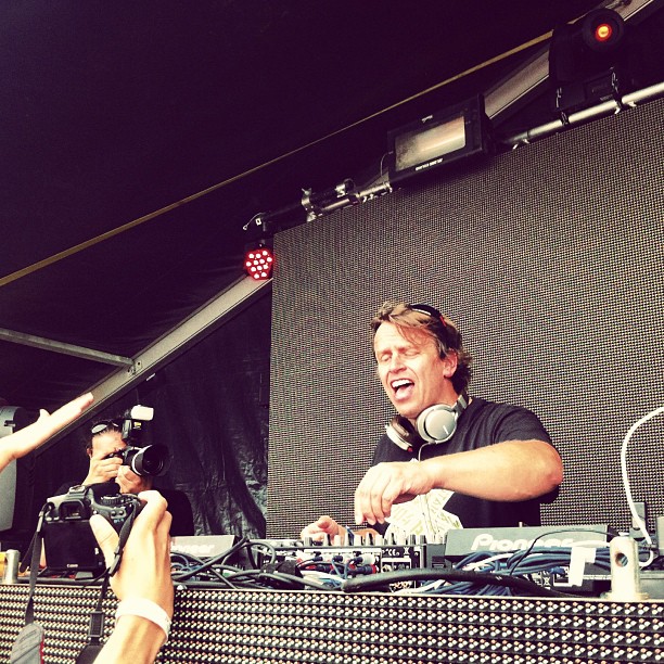 Chicane, Nick Bracegirdle, laughing at a DJ mixer with headphones round his neck while a photographer shoots from the side of the stage