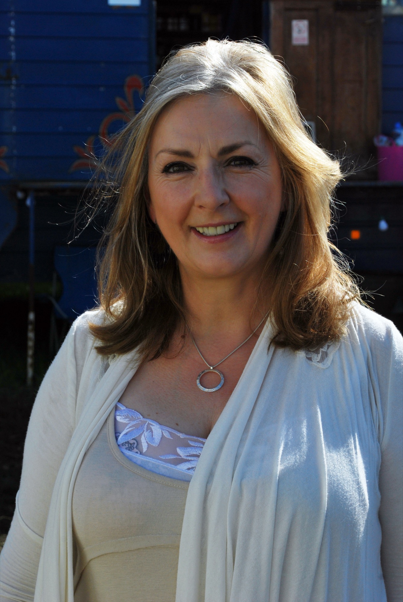 Moya Brennan smiling in a cream cardigan and silver pendant in front of a painted blue wooden wagon