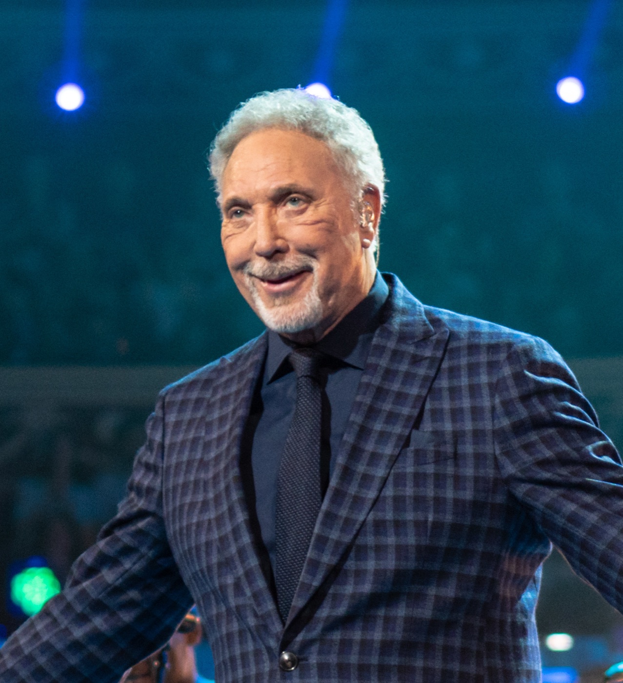 Tom Jones, white-haired and smiling, in a blue checked suit and dark tie on a stage under blue lights