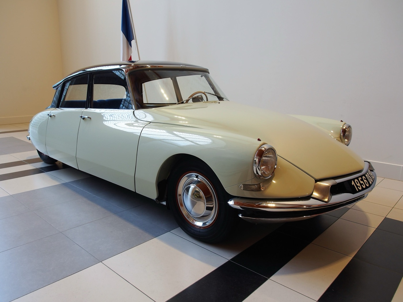 Front three-quarter view of a cream 1956 Citroën DS 19 with a dark roof, chrome bumper and round headlamps, in a museum hall beside a French flag