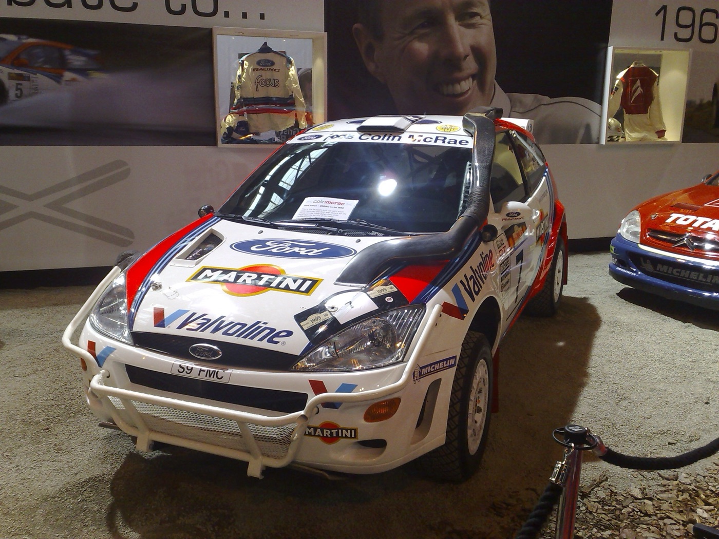 White Ford Focus WRC in Martini and Valvoline colours with a raised air snorkel and bull bar, under a large photograph of McRae smiling