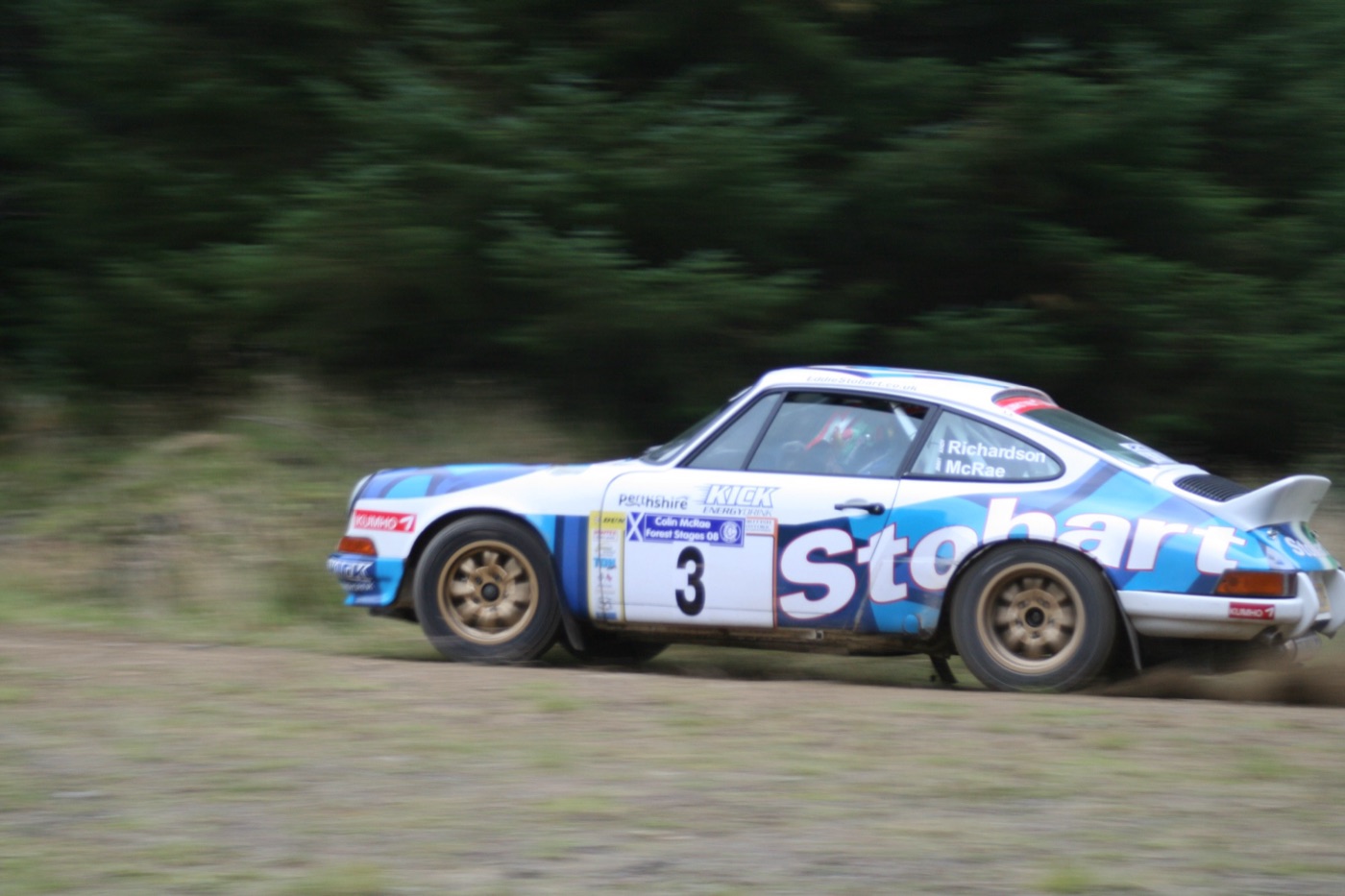 White and blue Porsche 911 rally car, number 3, sliding along a gravel forest track with the names Richardson and McRae on the side window