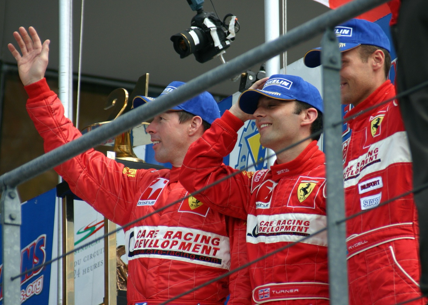 Colin McRae waving from the podium in a red Prodrive Ferrari race suit and blue cap, beside Darren Turner and Rickard Rydell