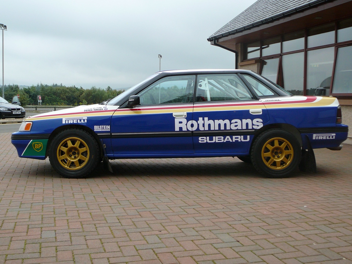 Blue, white, red and gold Subaru Legacy rally saloon in profile, with gold wheels and the names Colin McRae and Derek Ringer on the front wing