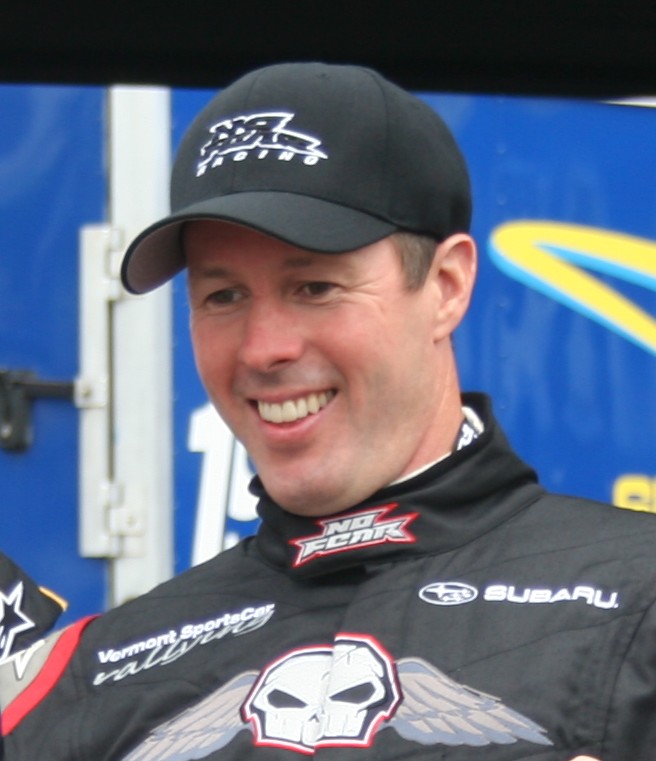 Head and shoulders of Colin McRae smiling, in a black cap and black race suit with Subaru and No Fear logos