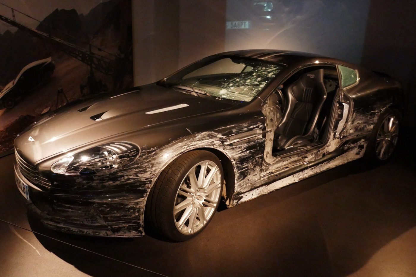 Battered grey Aston Martin DBS with scraped paint down its side, a missing door and a windscreen cracked by bullet holes, lit on a dark stand