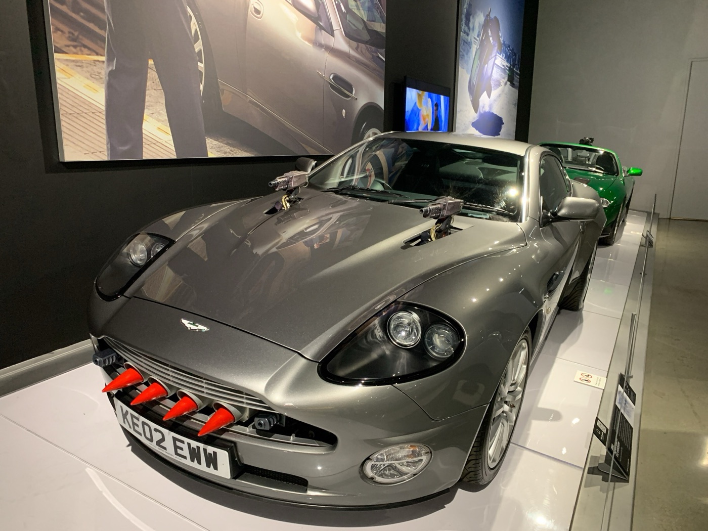 Silver Aston Martin V12 Vanquish with bonnet-mounted guns and red-tipped grille rockets, registration KE02 EWW, with a green Jaguar XKR parked behind