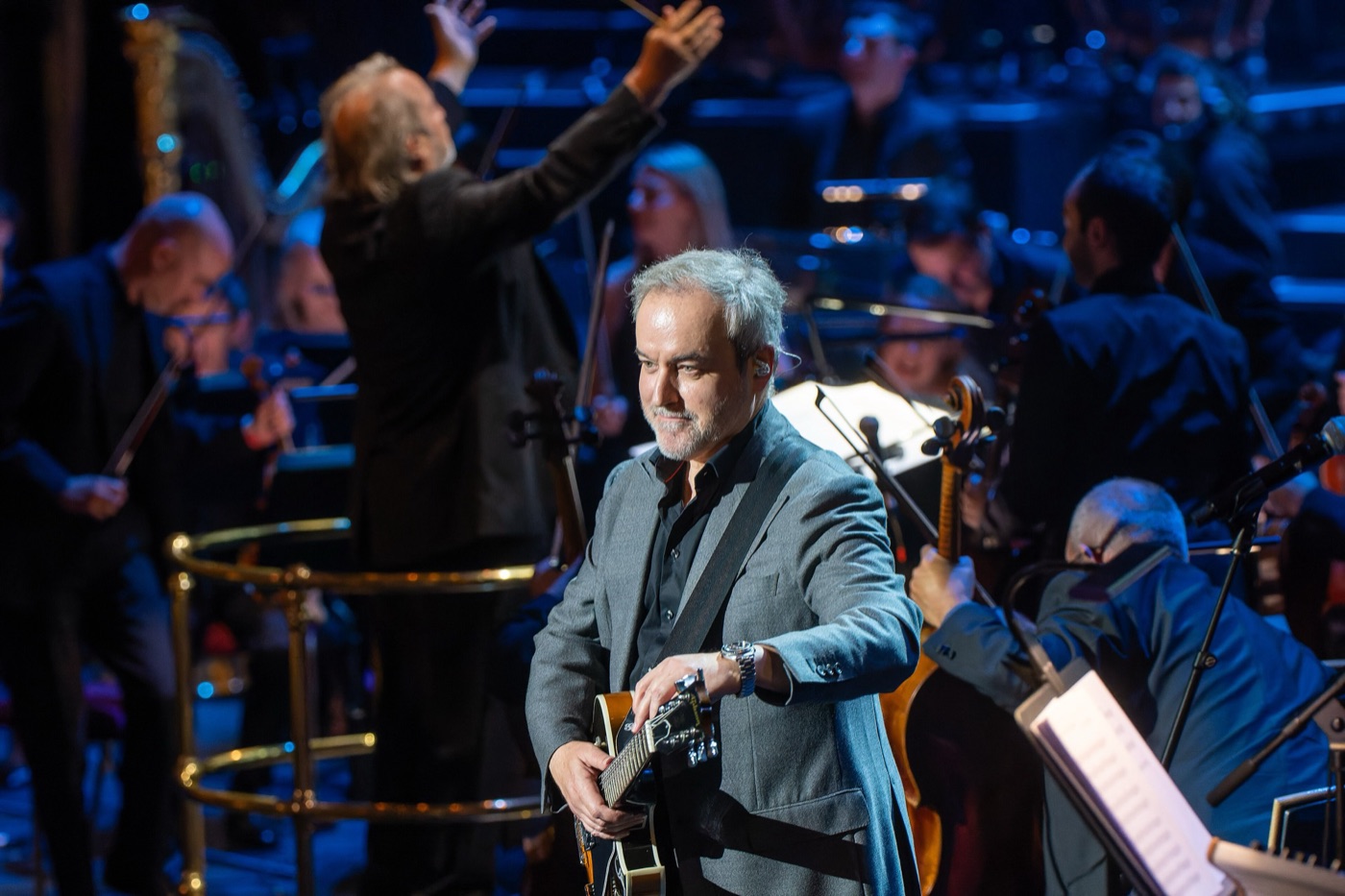 David Arnold in a grey jacket holding an electric guitar on stage in front of an orchestra and a conductor with raised arms
