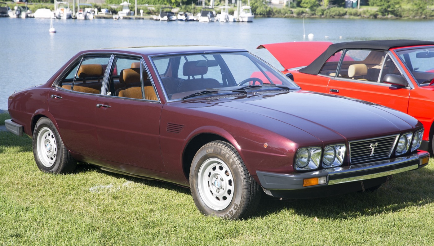 Dark red De Tomaso Deauville four-door saloon with quad headlamps, parked on grass by a marina