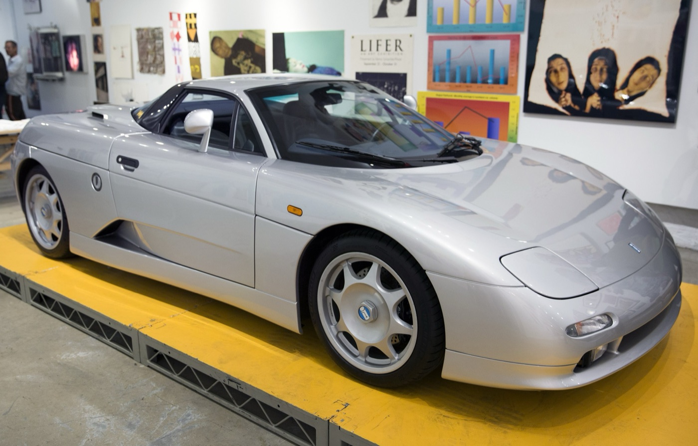 Silver De Tomaso Guarà coupé with round headlamp covers and five-spoke wheels, displayed on a yellow platform in a gallery