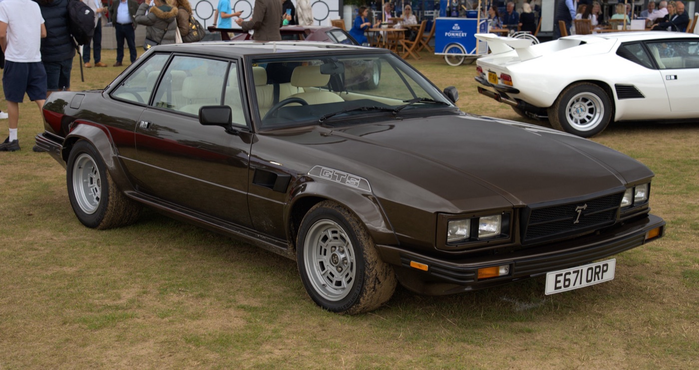 Brown De Tomaso Longchamp GTS coupé with flared arches and GTS lettering on the wing, on a showground with a white De Tomaso Pantera behind