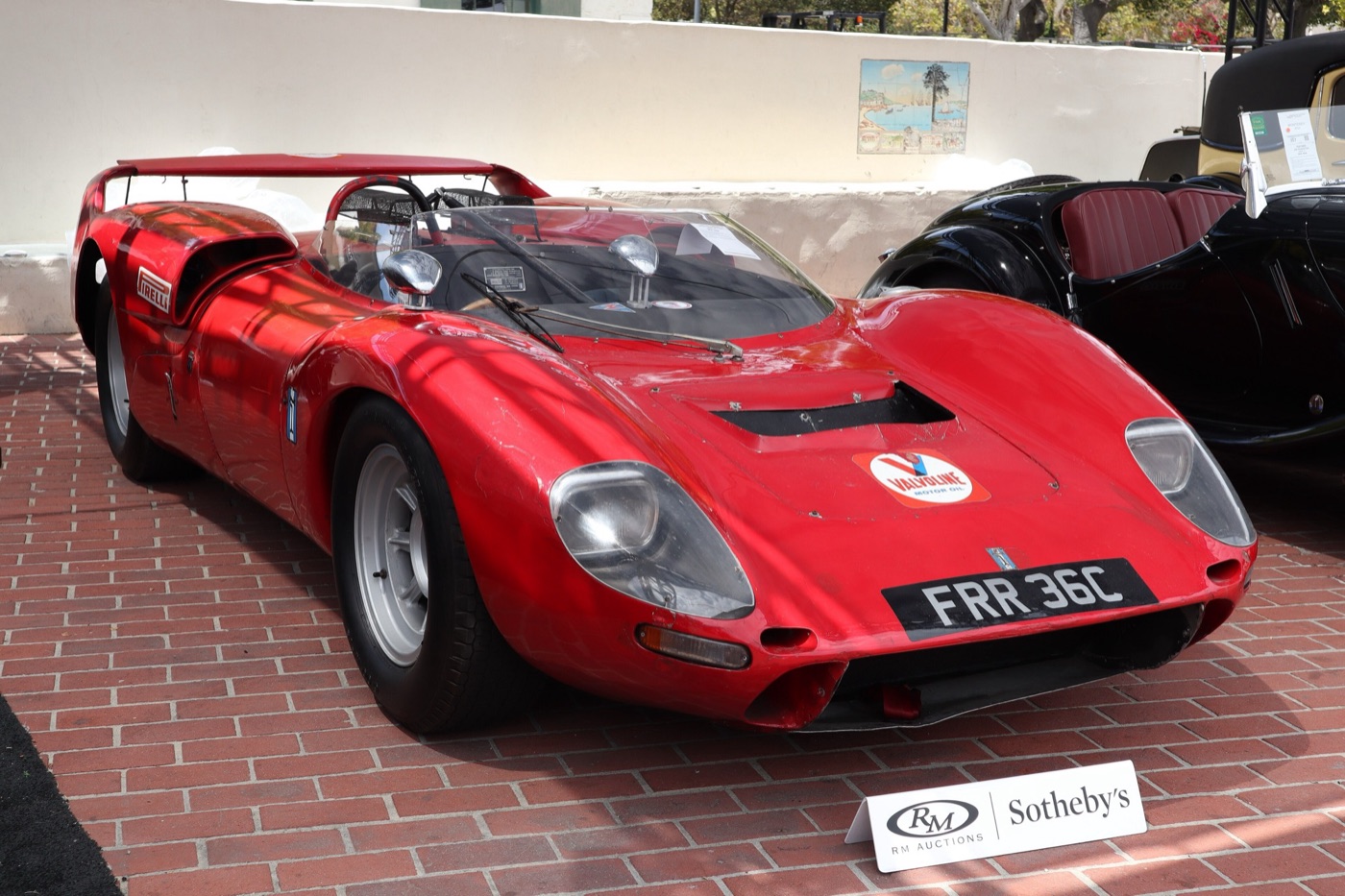 Red open De Tomaso Sport 5000 racing car with a high rear wing and a British number plate, on display at an auction