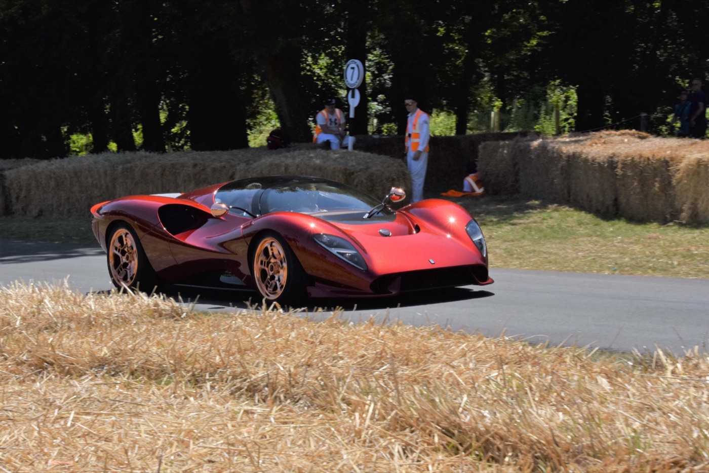 Red De Tomaso P72 with copper-coloured wheels driving up the hill at Goodwood between straw bales, with marshals in orange vests behind