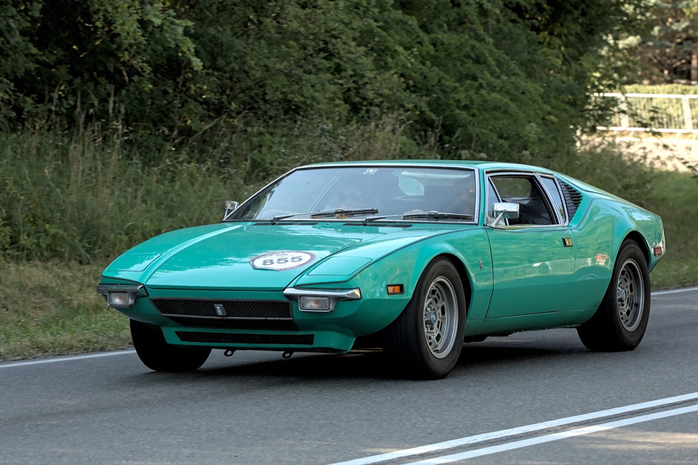 Green 1971 De Tomaso Pantera with a rally number on the bonnet, driving along a tree-lined road