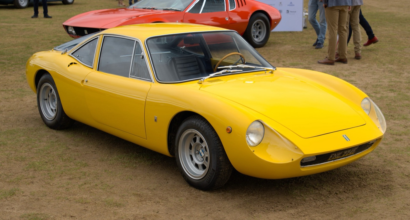 Yellow De Tomaso Vallelunga coupé with a long glass rear hatch, parked on grass at a concours with a red De Tomaso Mangusta behind
