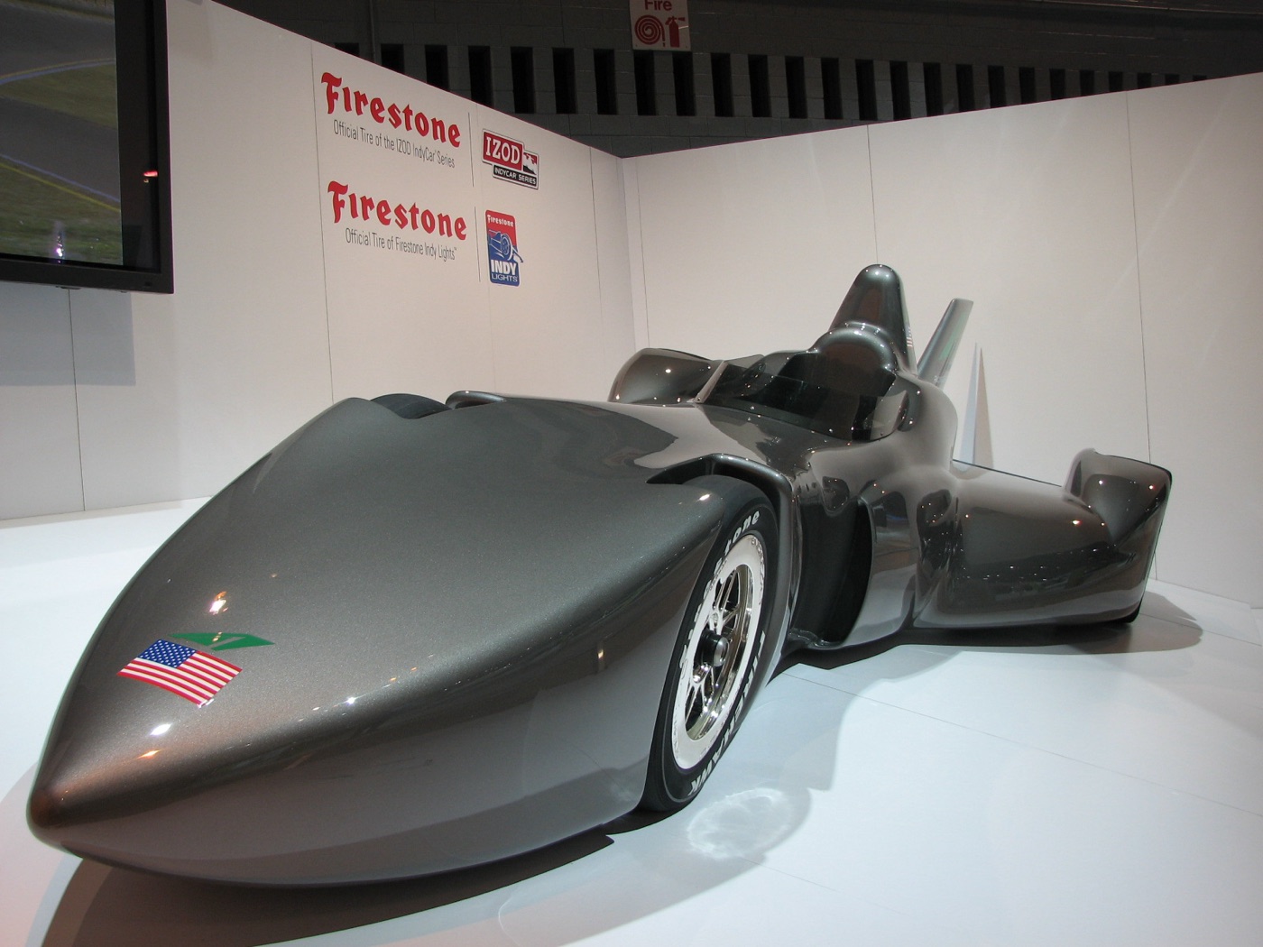 Grey DeltaWing IndyCar concept show car with a long pointed nose and narrow front wheels on a white stand