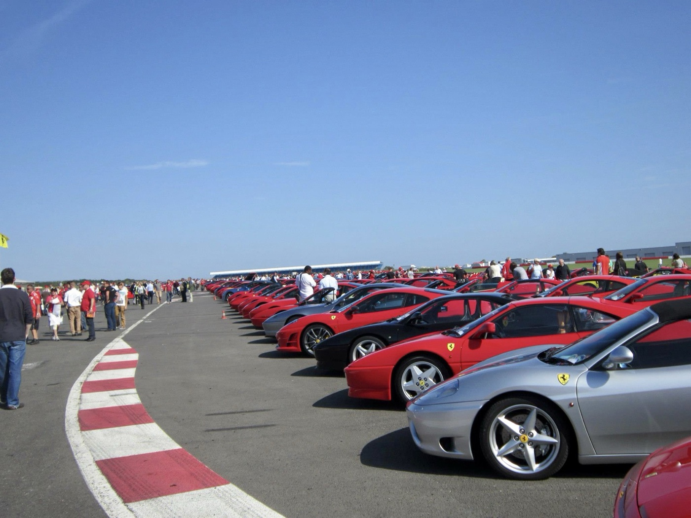 A long line of red, black and silver Ferraris queued on the track at Silverstone beside a red and white kerb, with spectators on the left