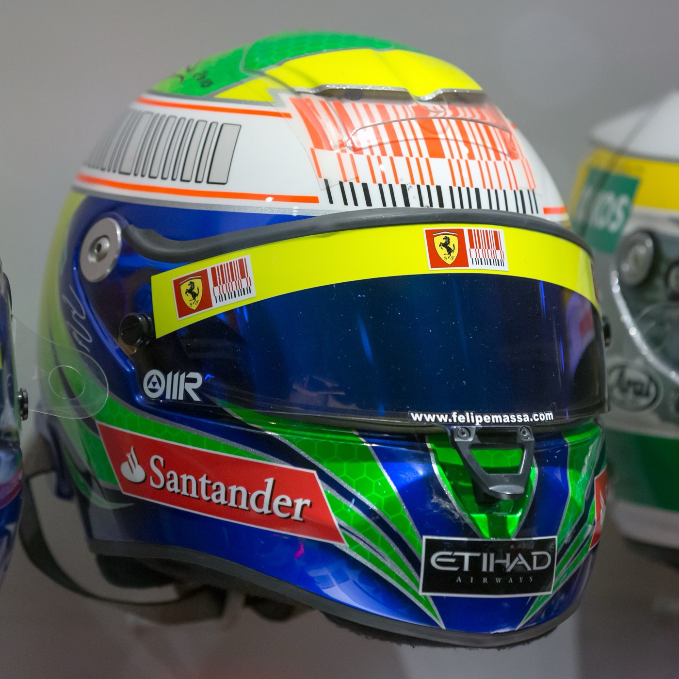 Felipe Massa’s blue, yellow and green 2010 racing helmet with Ferrari prancing horse stickers and a yellow strip across the top of the visor, on display in a museum
