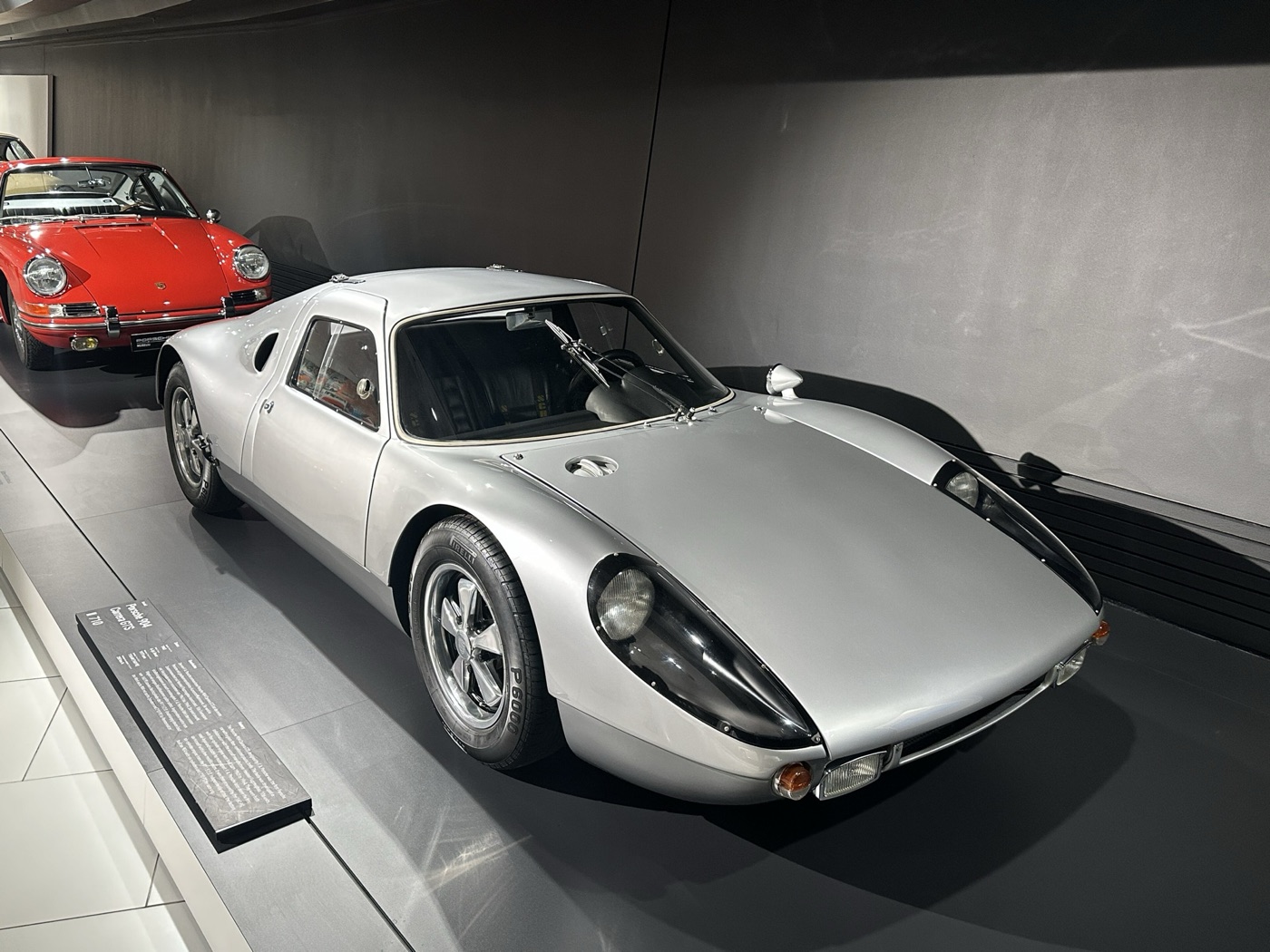 Silver Porsche 904 Carrera GTS with headlamps behind clear covers, parked in front of a red early 911 in a museum
