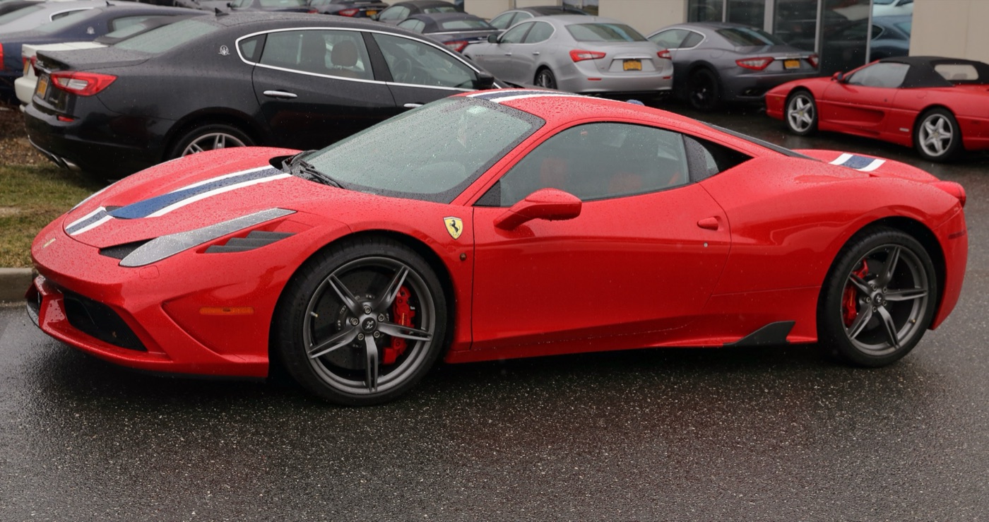 Red Ferrari 458 Speciale with a blue and white stripe and dark five-spoke wheels parked on a wet forecourt