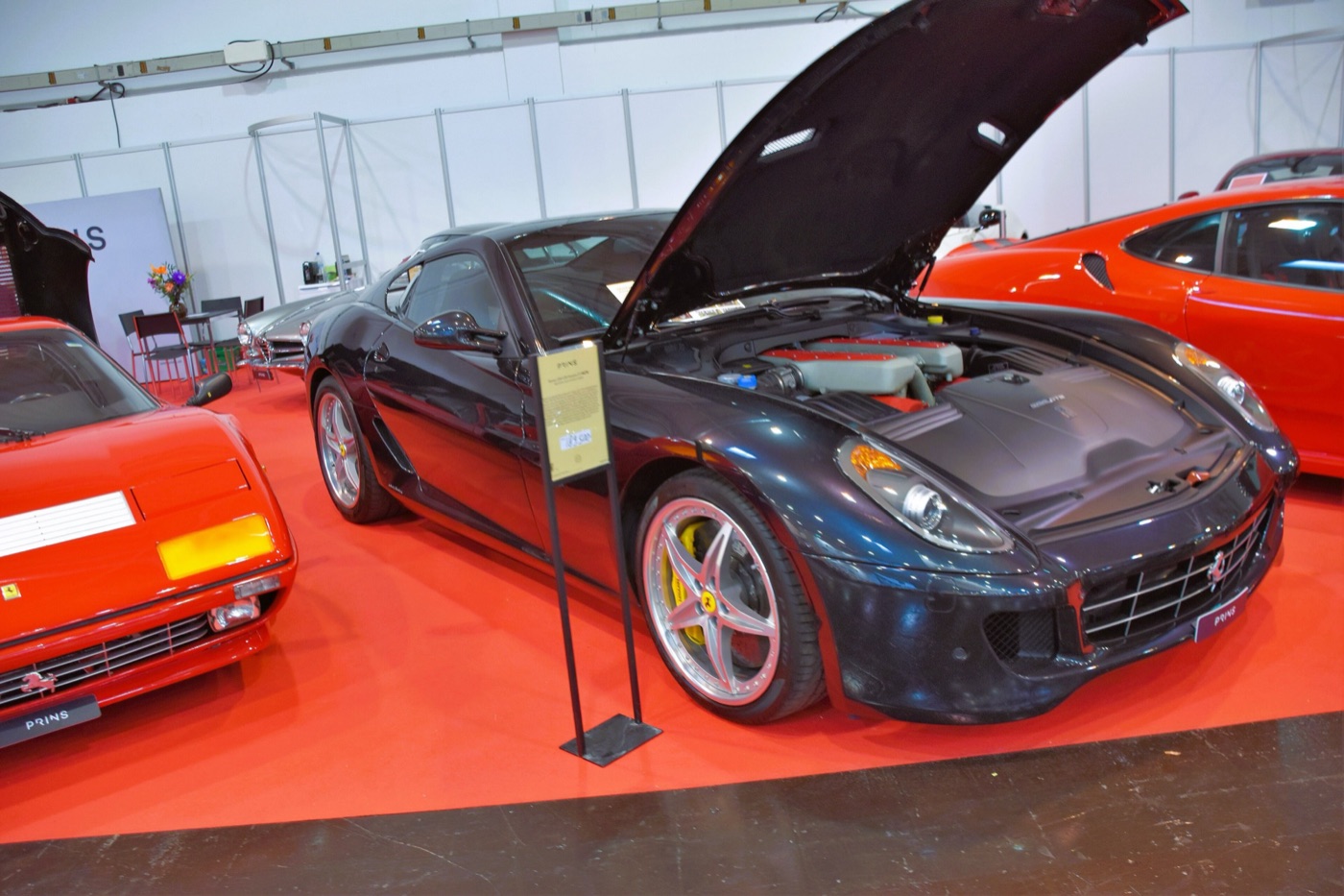 Dark blue Ferrari 599 GTB Fiorano with its bonnet open on a red show stand between other Ferraris