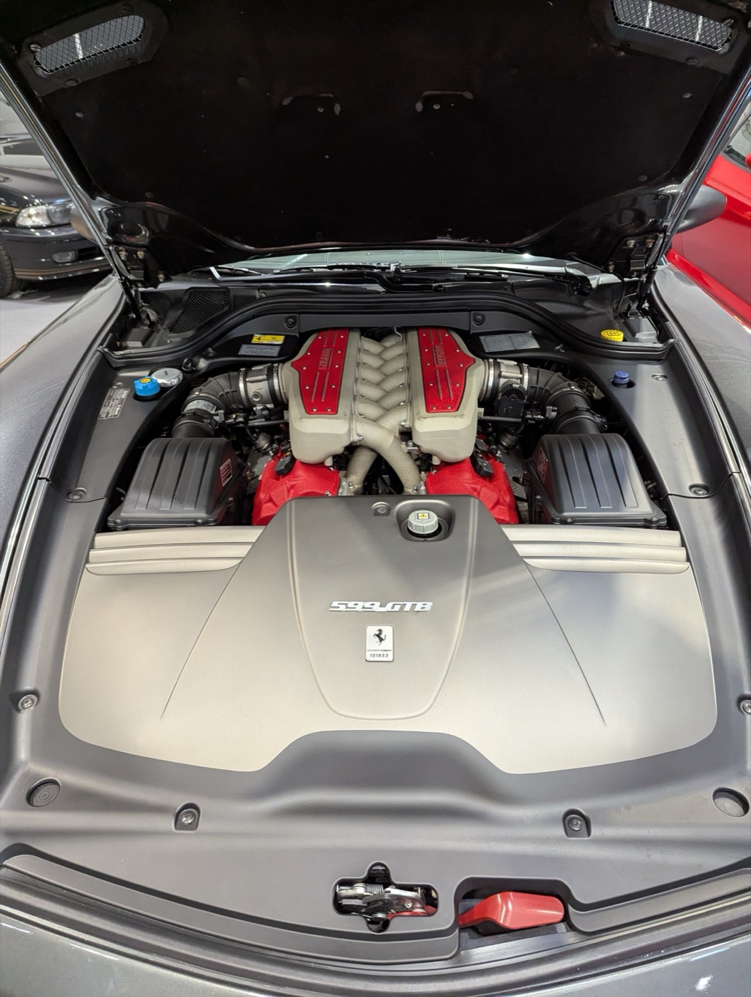 Engine bay of a Ferrari 599 GTB with the red V12 intake manifolds behind a grey cover marked 599 GTB