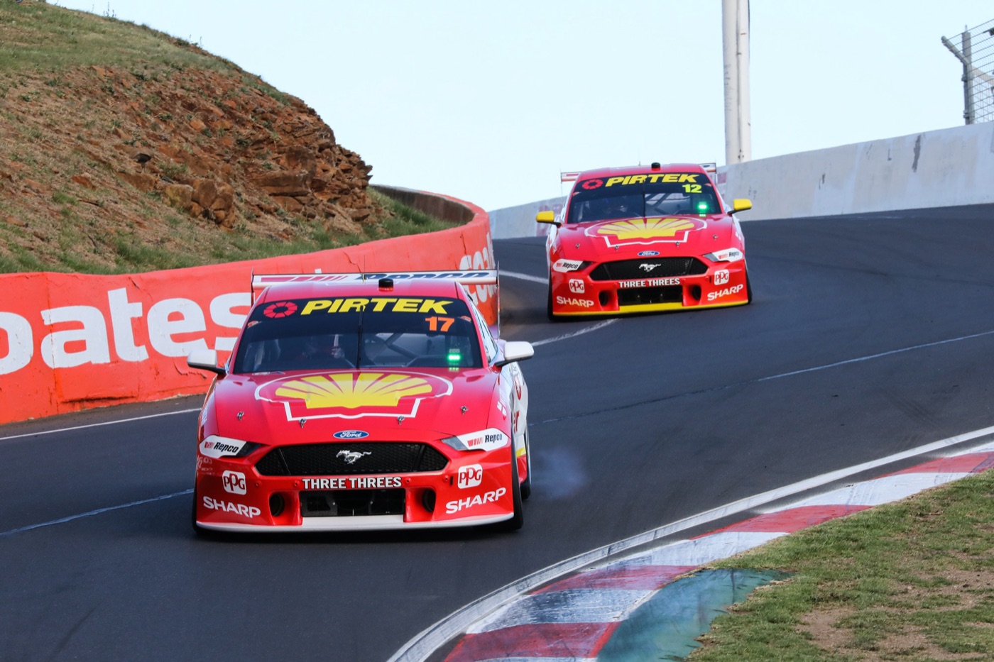 Two red and yellow Shell-liveried Mustang Supercars, numbers 17 and 12, climbing a steep corner beside a rock face and a concrete wall