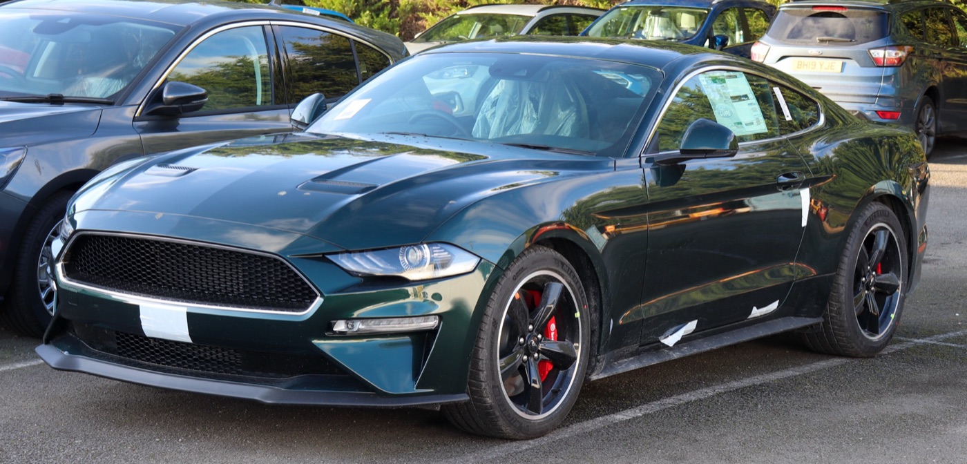 Dark Highland Green Mustang Bullitt with black five-spoke wheels and red brake calipers, still with protective tape on the bumper, in a car park