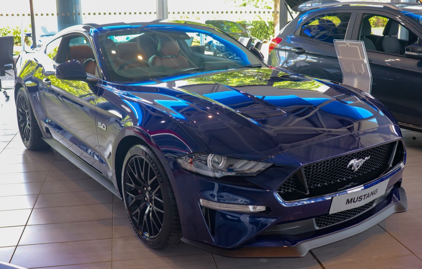 Dark blue facelifted Mustang GT with black wheels and bonnet vents, tan leather seats visible, in a Ford showroom