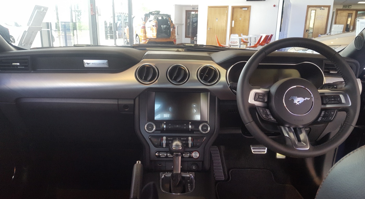 Right-hand-drive Mustang dashboard with a three-spoke steering wheel, three round air vents, a central touchscreen and an automatic gear selector, in a Ford showroom