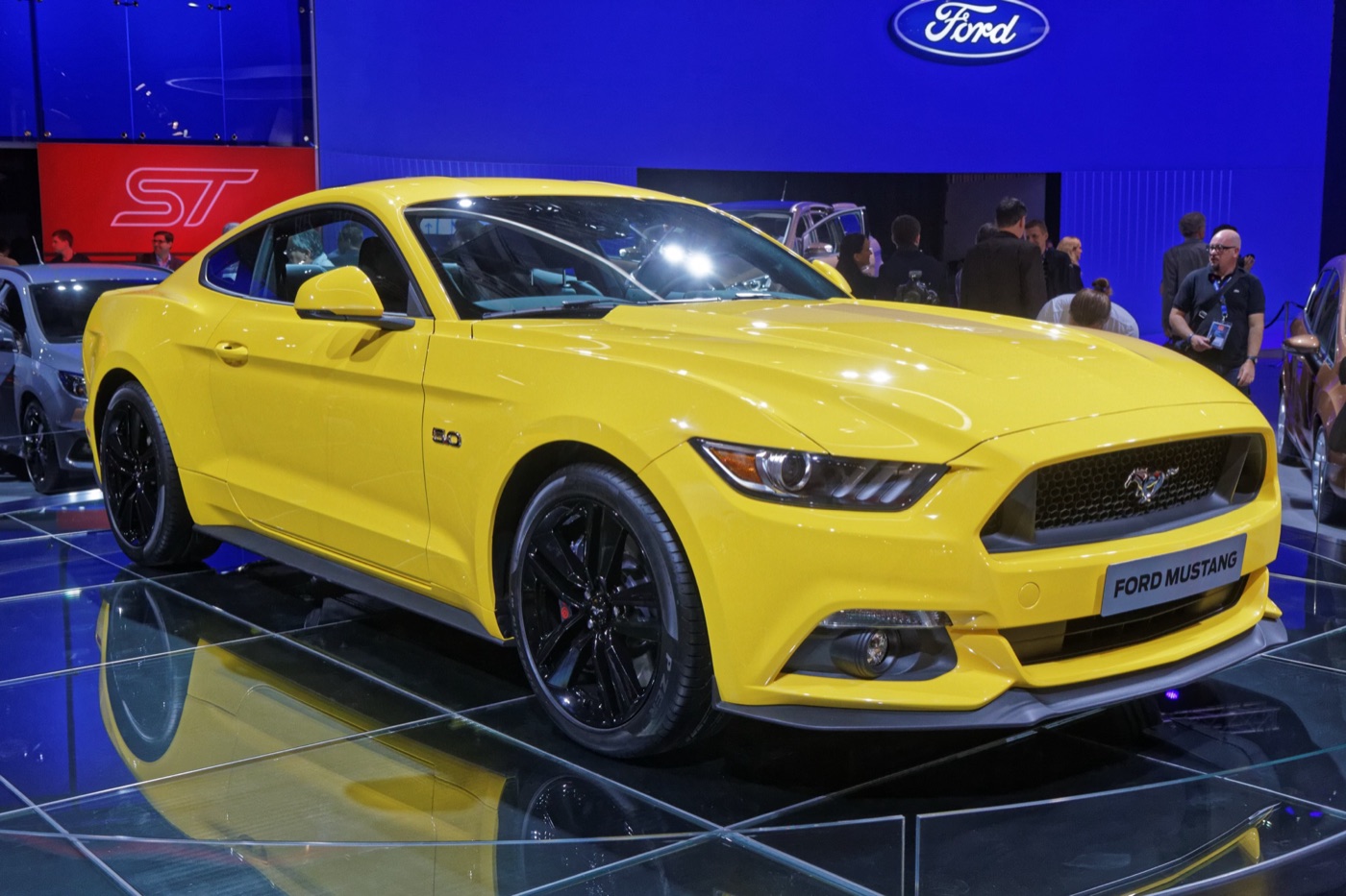 Yellow Mustang fastback with black wheels and a 5.0 badge on the Ford stand at the Paris motor show, people behind