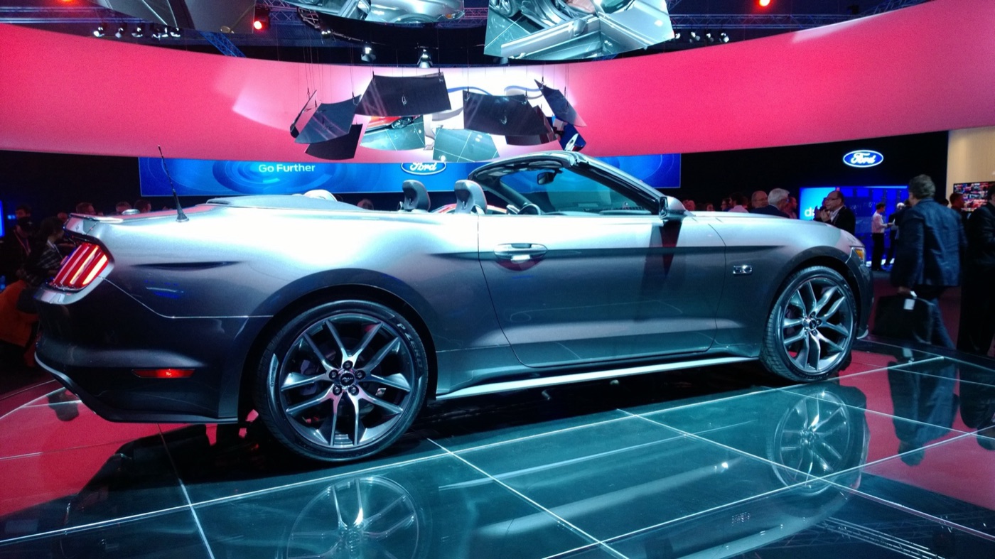 Grey Mustang convertible with its roof down on a glass turntable under pink and blue stage lighting, with Ford Go Further banners behind