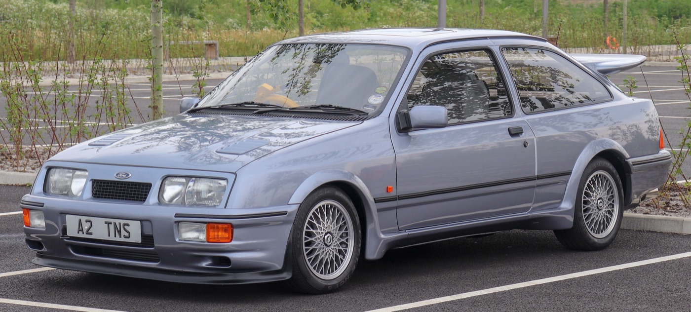 Moonstone Blue three-door Ford Sierra RS Cosworth seen from the front three-quarter, with its front air dam, spoked alloy wheels and rear wing, in a car park