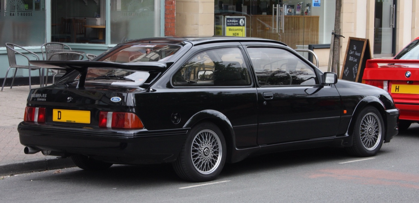 Rear three-quarter view of a black three-door Ford Sierra RS Cosworth parked at a kerb, showing its tall rear wing on the tailgate and multi-spoke wheels