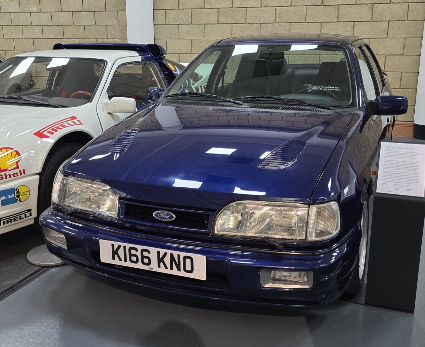 Dark blue four-door Ford Sierra Sapphire Cosworth 4x4 with two bonnet louvres, displayed indoors beside a white rally car
