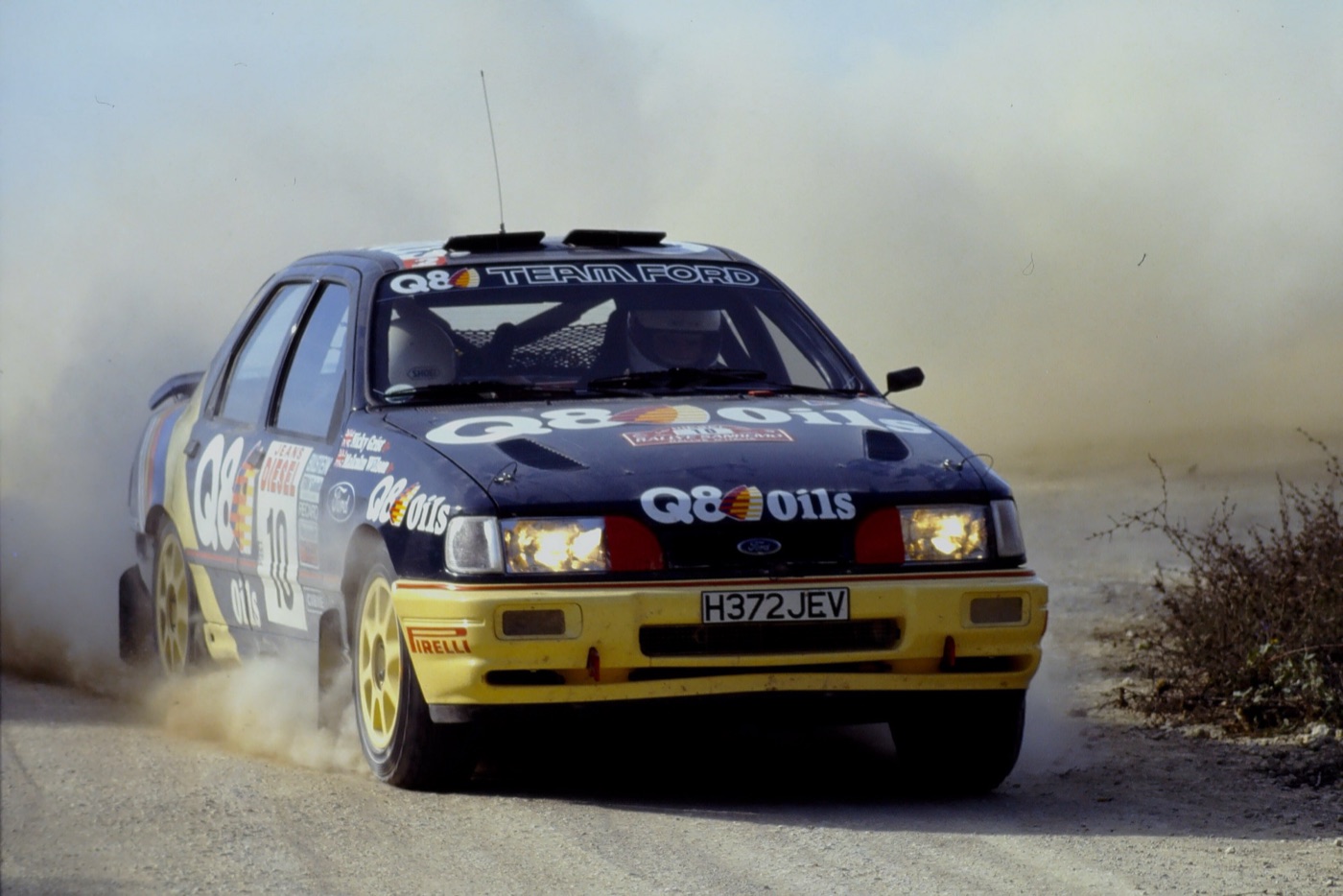 Blue and yellow Q8 Team Ford Sierra Cosworth 4x4 rally car, number 10, sliding on a dusty gravel road