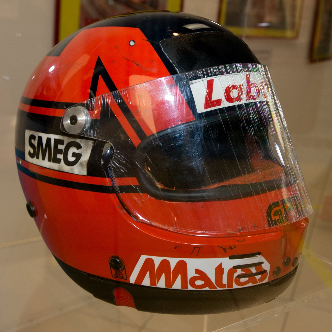 Red and black racing helmet with a V shape on the side and Smeg, Labatt and Matras stickers, in a display case