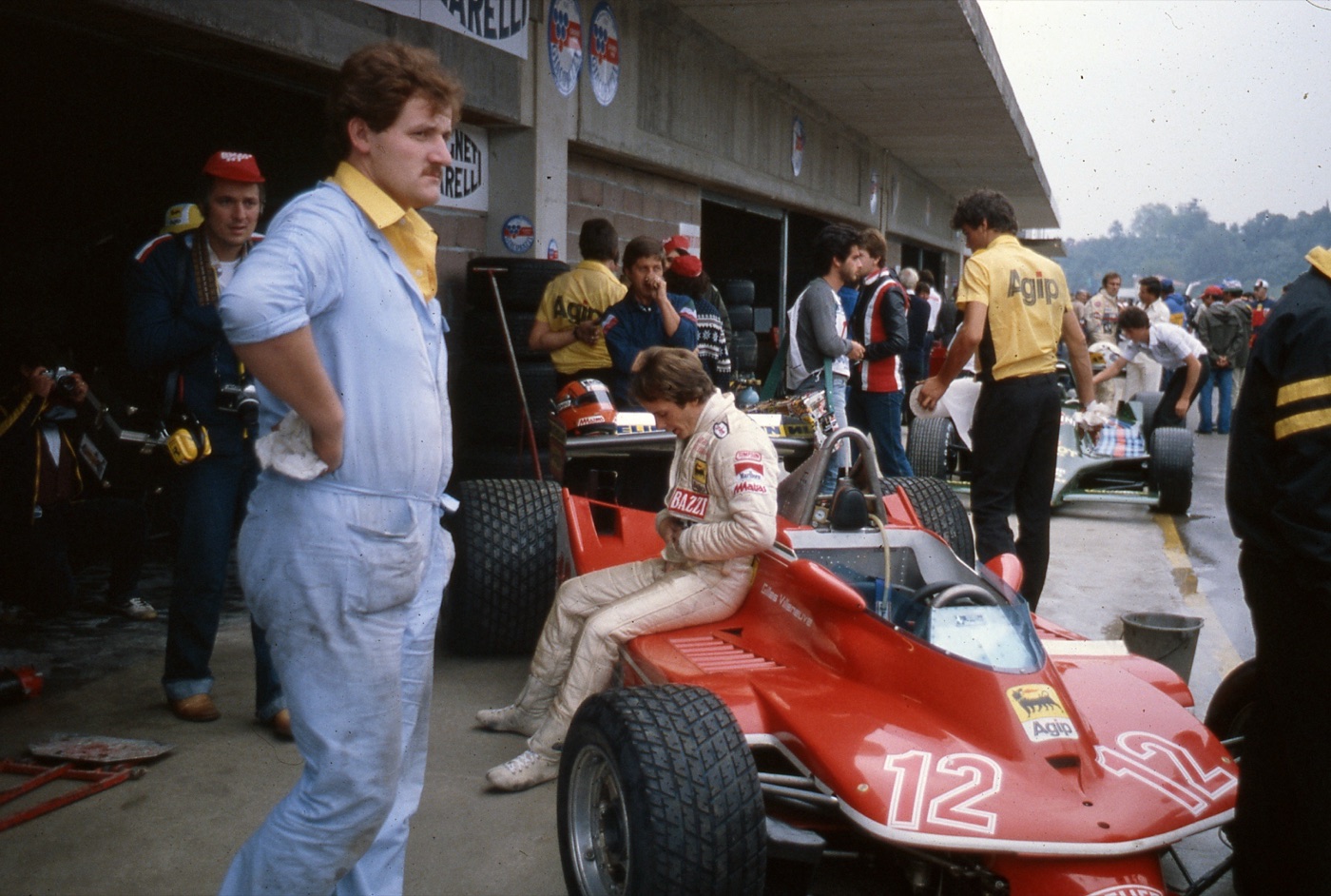 Gilles Villeneuve in white overalls sitting on the side of his red Ferrari number 12 in the Imola pit lane, mechanics and photographers around him