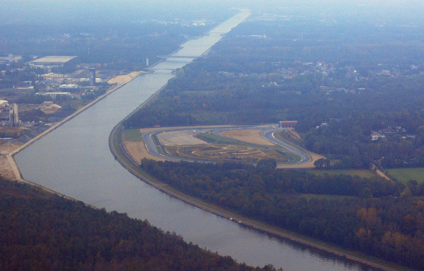 Aerial view of a curving canal with the western loop of a racing circuit on its inside bank, surrounded by woodland