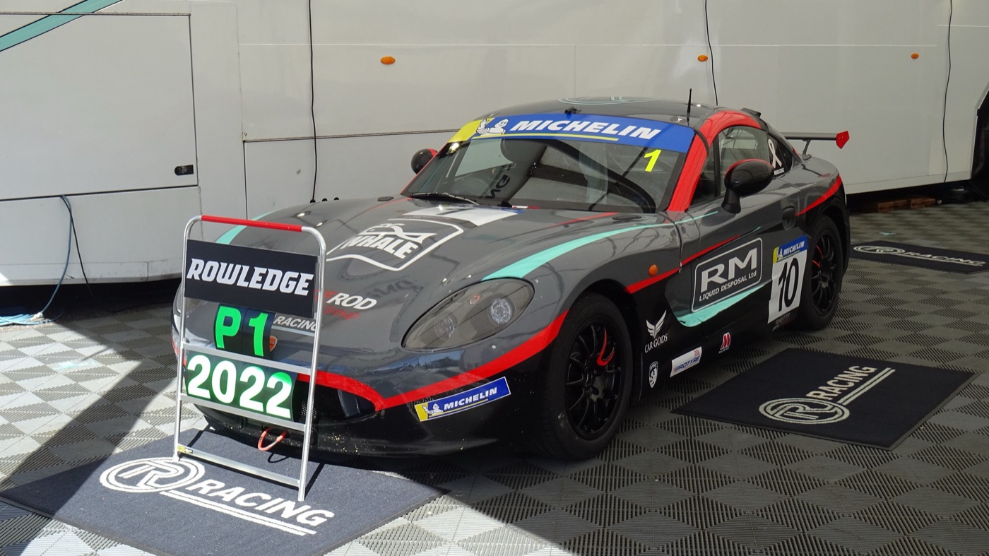 Grey, red and turquoise Ginetta G40 Junior race car in a team awning behind a P1 2022 board