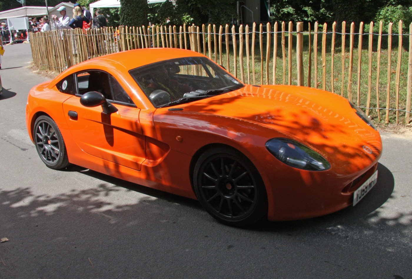 Orange Ginetta G40 coupé with black alloy wheels parked by a wooden fence in dappled shade