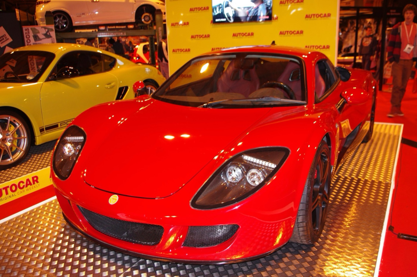 Red Ginetta G60 mid-engined coupé on a show stand in front of a yellow Autocar backdrop