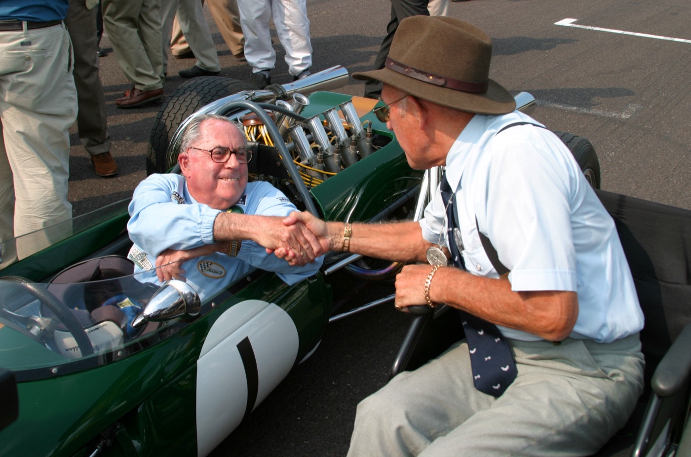 Jack Brabham, seated in his green Brabham BT19 Formula 1 car, shakes hands with Stirling Moss in a brown trilby and tie at the 2004 Goodwood Revival